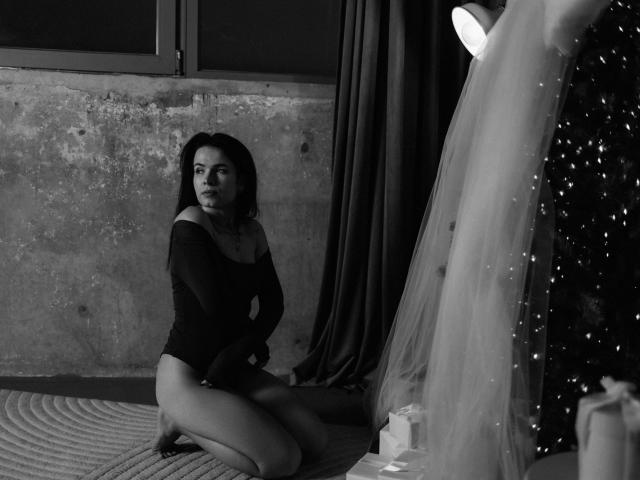 JasmineKouch, black-haired, off-shoulder top, kneeling, showing ass, on rug, next to window, with curtains and hanging light, in black-and-white photo.