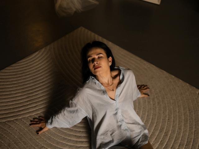 JasmineKouch, with black hair, is lying on a textured rug, wearing a light blue shirt, eyes closed, arms spread, and a necklace, in a dimly lit room.