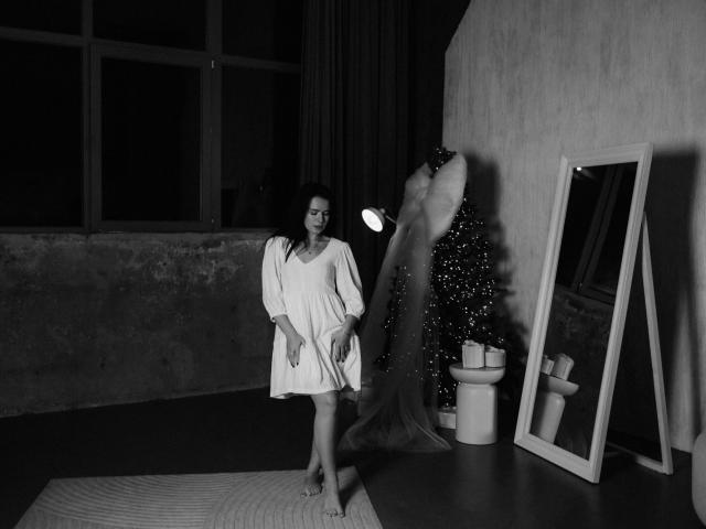 JasmineKouch in a white dress stands barefoot, holding a flashlight, next to a mirror and Christmas tree, with a standing lamp and wooden floor in a dark room.
