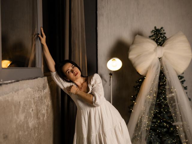 JasmineKouch, in a white dress, leans against a window, her arm raised, with a lit lamp and decorated Christmas tree with a giant white bow in the background.