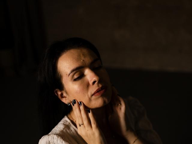 JasmineKouch has dark hair, closed eyes, and a slight grimace, touching her neck with black-manicured hands. She's wearing a white, textured top, in dim, moody lighting.