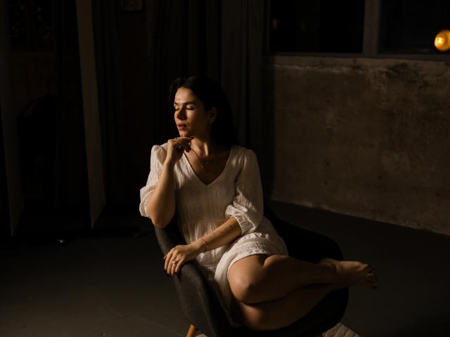 JasmineKouch with medium tits, wearing a white dress, sitting on a black chair, legs crossed, dark hair, touching her chin, dimly lit, concrete wall, soft brown light.