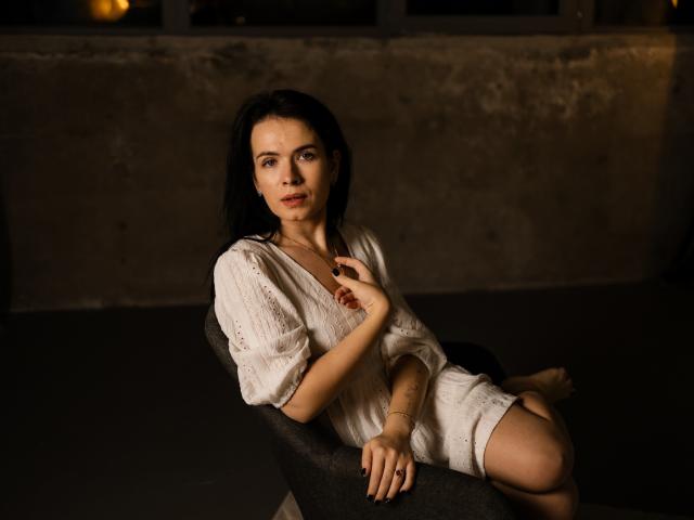 JasmineKouch with dark hair, white lace dress, sitting on black chair, hand on chest, soft lighting, bare legs, slight smile, dark background, subtle sexy, relaxed, attractive.