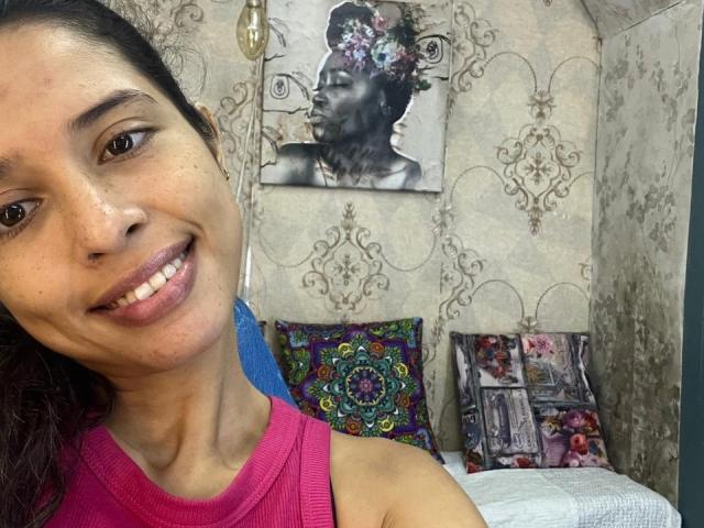 LunaVelvett has medium brown skin, dark brown eyes, and a small nose with a pink tank top, smiling with her teeth showing. She has a flower in her hair.