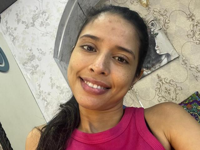 LunaVelvett, brown-eyed, smiling, tan-skinned, with black hair in a ponytail, wearing a pink tank top, showing her small, cute nose and slightly freckled face.