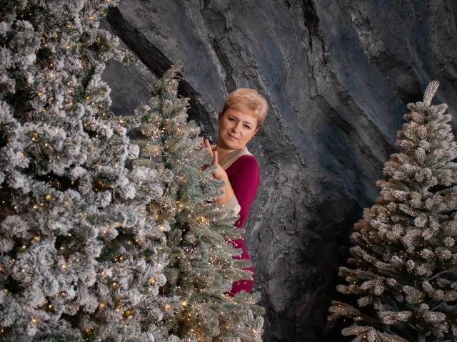 EmiliaGree with short blonde hair, wearing a red top, peeks between two frosted Christmas trees, smiling slightly, surrounded by twinkling lights and a rocky background.