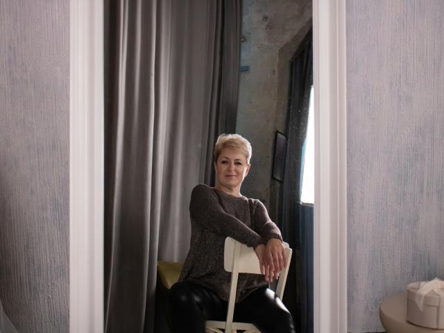 EmiliaGree with short blonde hair, gray sweater, black leather pants, sitting on white chair, hands on seat, slight smile, gray curtains, textured walls, natural light from right.
