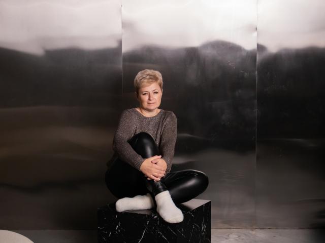 EmiliaGree sits cross-legged on a black marble block, wearing a gray sweater, black leather pants, white socks, and looking slightly annoyed. Her short blonde hair is visible.