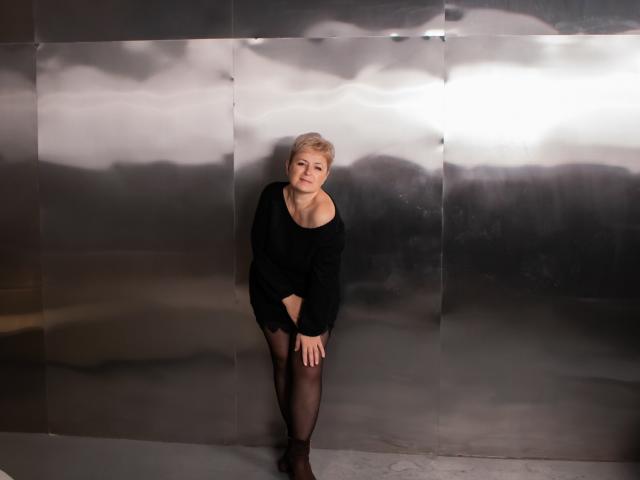 EmiliaGree with short blonde hair, wearing a black off-shoulder sweater and black sheer stockings, leaning against a shiny metallic wall, looking slightly downward with a neutral expression.