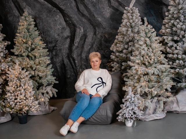 EmiliaGree with short blonde hair, white sweater, blue jeans, and white shoes, sitting on gray beanbag between two frosted Christmas trees, hands on knees, looking slightly annoyed.