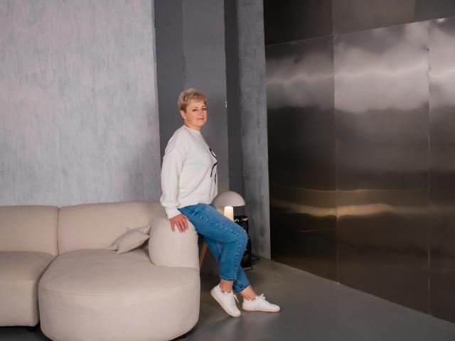 EmiliaGree with short blonde hair, white sweater, blue jeans, and white sneakers, sitting on a beige sectional sofa, looking to the right, in a modern, metallic room.
