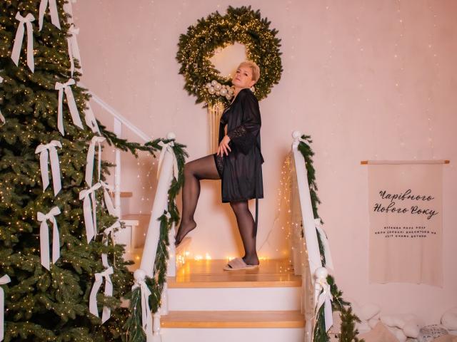 EmiliaGree with blonde hair, black lace dress, and sheer black stockings, posing on staircase, one leg up, showing her ass, standing in front of a Christmas wreath.
