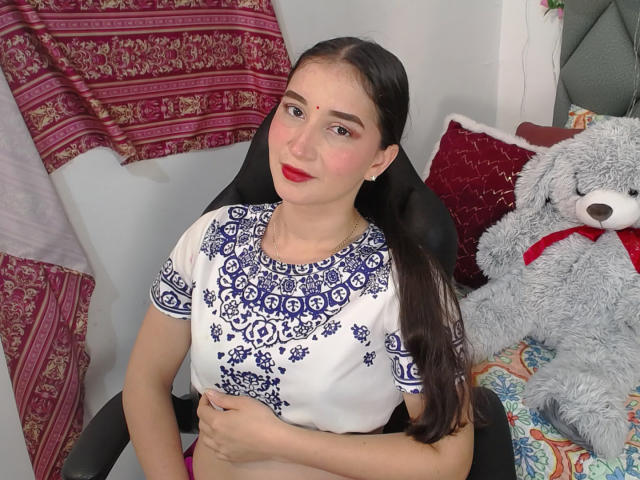 JalimaMona with long dark hair, red lipstick, white top with blue patterns, sitting in black chair, hand on belly, gray stuffed teddy with red ribbon on colorful bed.