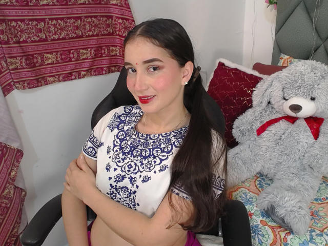 JalimaMona with long dark hair, red lipstick, and blue patterned top, arms crossed, sitting on chair, next to gray teddy bear with red bow, on colorful bed.
