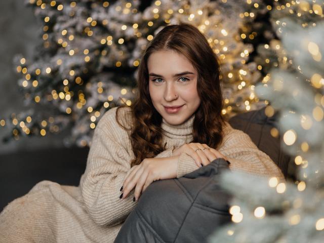 LauraHlot is a young woman with wavy brown hair and light skin, sitting on a gray cushion. She's wearing a beige knit turtleneck. Her hands rest on the cushion, and she has a subtle smile. The background is blurred with warm, golden bokeh lights from Christmas trees. The texture of her sweater is visible, and her nails are painted black. The composition focuses on her relaxed, comfortable posture. The light is soft, highlighting her facial features and creating a cozy atmosphere.
