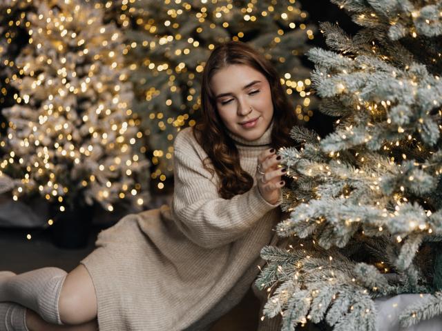 LauraHlot with curly brown hair, smiling, wearing a beige knit sweater, touching a frosty Christmas tree, surrounded by warm, glowing lights. Her ass is partly visible.