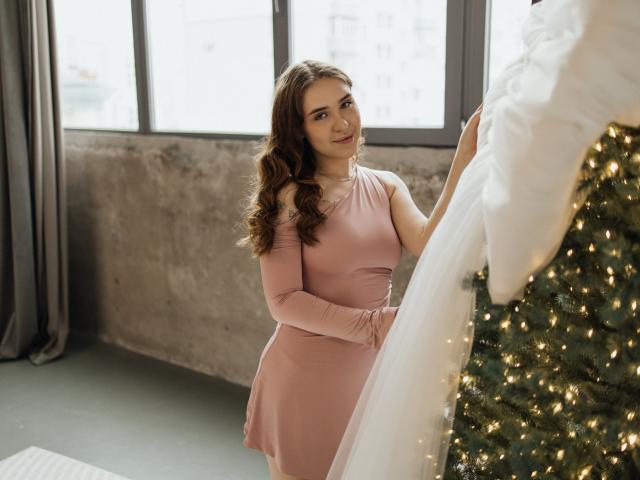 LauraHlot with wavy brown hair, pink dress, and cute smile, holding white fabric, standing next to a decorated Christmas tree with warm lights.