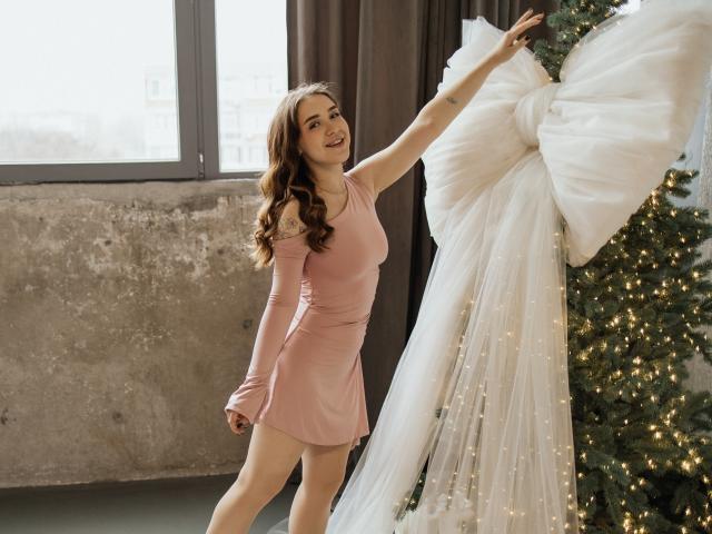 LauraHlot with wavy brown hair, pink dress, and small tits, touching a white, giant, fluffy bow on a Christmas tree with warm lights in a sunlit room.