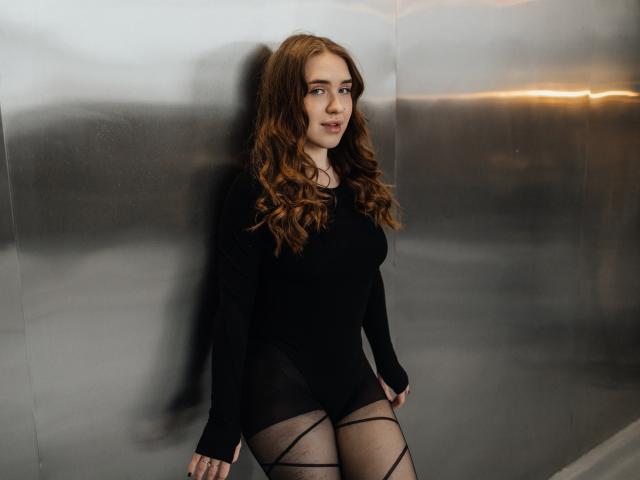 LauraHlot with wavy red hair, black long-sleeve top, and black mesh pantyhose, standing against a shiny metal wall, smiling subtly, arms relaxed. Boobs, ass, pussy not visible.