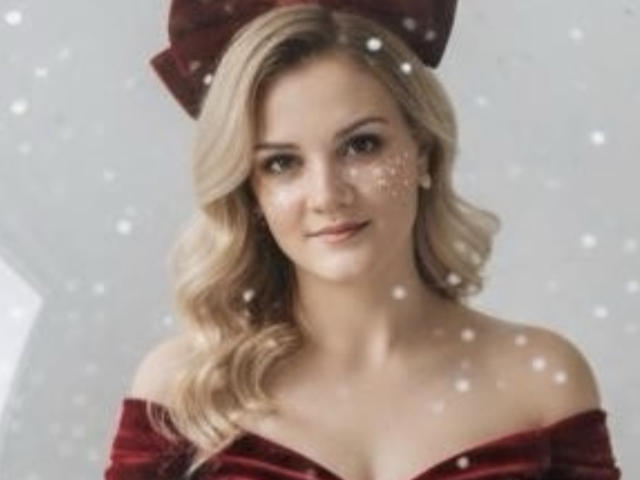 SarahFontain with blonde wavy hair, red velvet off-shoulder dress, and matching hat, small sparkly face makeup, looking at camera, subtle smile, white snowflakes in background.