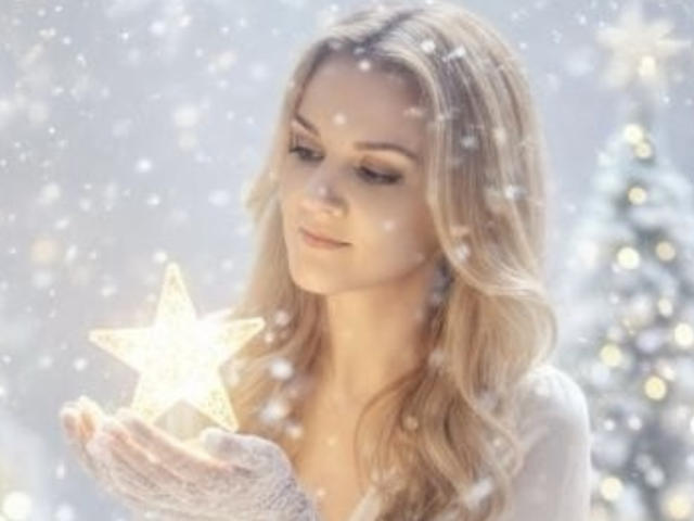 Sarah Fontain mit langem, blondem Haar, die einen leuchtenden Stern in ihrer mit weißen Handschuhen bedeckten Hand hält, Schneeflocken fallen, sie trägt ein weißes Oberteil, der Weihnachtsbaum ist im Hintergrund unscharf..