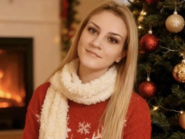 SarahFontain with long blonde hair, red sweater, white fluffy scarf, and Christmas tree with red and gold ornaments, smiling softly, standing near a lit fireplace.