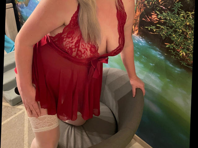 HotLeyla86-sex, wearing red lace lingerie with sheer fabric, white thigh-high stockings, leaning on gray chair, showing large boobs, blonde hair, red bow, in cozy indoor setting.
