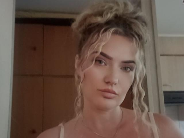 CarmenAutumn has curly blonde hair in a messy bun, wearing a white tank top, with a nose ring and subtle makeup, looking serious with pouty lips.