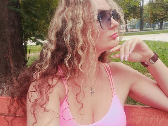 CarmenAutumn with curly blonde hair, wearing a pink sports bra, showing her tits, sunglasses, and a cross necklace, resting her chin on her hand, wristwatch on.