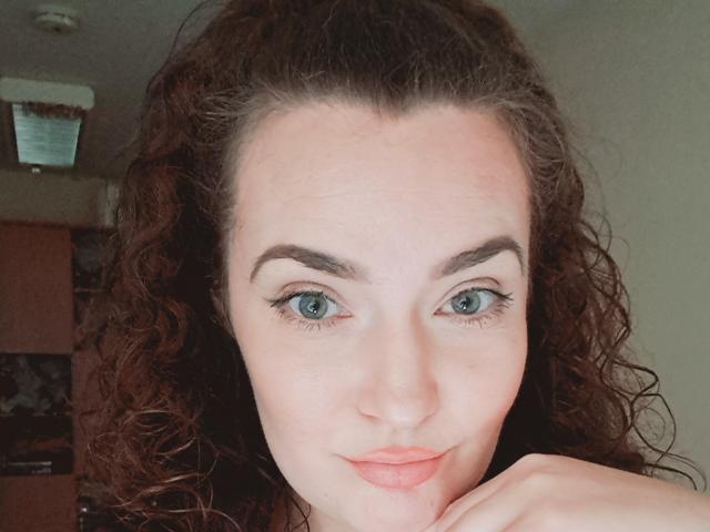 CarmenAutumn has curly brown hair, blue eyes, and a slight smile. She's resting her chin on her hand, looking directly at the camera. Her tits are not visible.