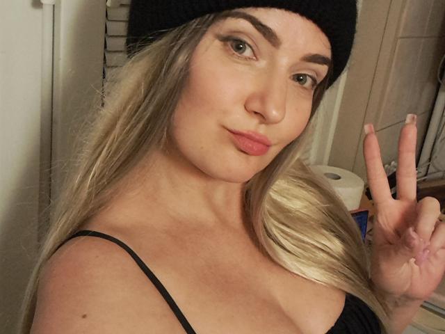 CarmenAutumn with blonde hair, black beanie, and black top, showing off her boobs with a cute pout and making a peace sign, in a bathroom selfie.