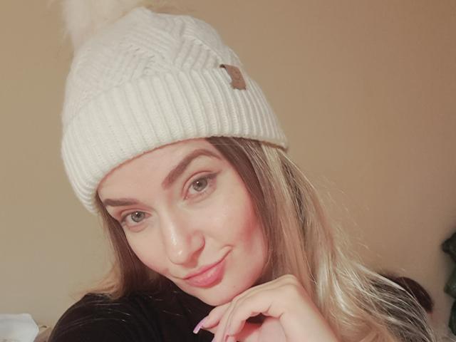 CarmenAutumn with blonde hair, wearing a white knit beanie, black top, and a slight smile, looking at the camera with green eyes, pink cheeks, and a cute, sexy look.