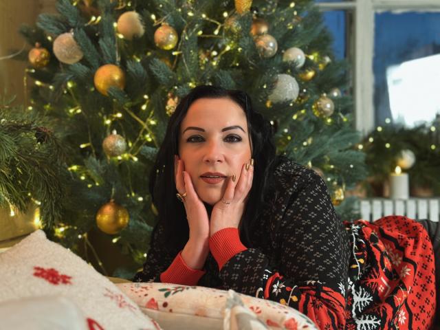 ElizabethSo with black hair, hands on cheeks, wearing a black and red patterned sweater, lying in front of a Christmas tree with gold ornaments and lights, looking cute.