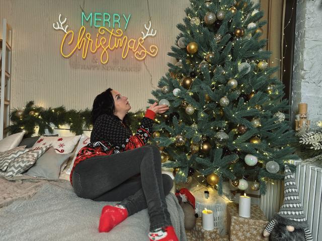 ElizabethSo with dark hair, wearing a black and red Christmas sweater, black pants, and red socks, sitting on a couch, reaching up to decorate a Christmas tree.