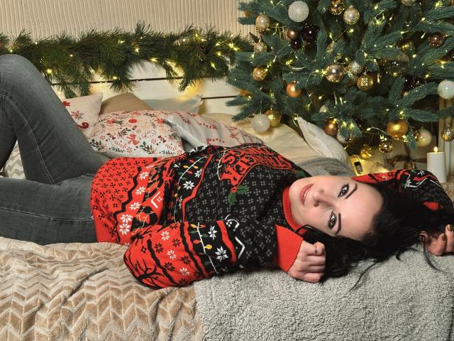 ElizabethSo is lying on a bed, wearing a red and black Christmas sweater, grey jeans, and smiling at the camera with her arm raised. She's next to a decorated tree.