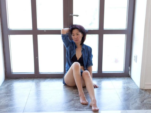SandraMiy with short black hair, sitting on marble floor, wearing an unbuttoned blue shirt, black bra, and no pants, legs crossed, hand in hair, barefoot, looking at camera.