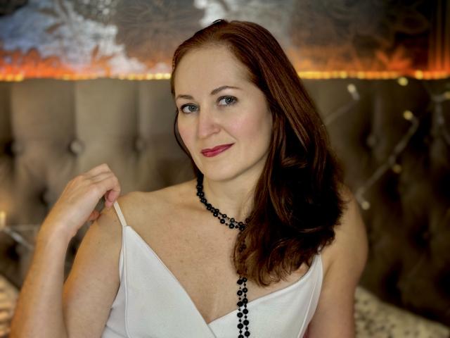JasmineeSky with red hair, fair skin, and pink lipstick, holding a black bead necklace, wearing a white strap dress, smiling gently, showing her small tits.