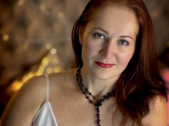 JasmineeSky with red hair, wearing a white dress and black bead necklace, smiling softly at the camera. Her tits are modest, with fair skin and pink lipstick.