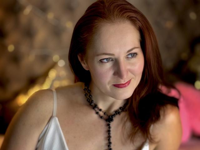 JasmineeSky with red hair, blue eyes, and red lipstick, wearing a black bead necklace, white spaghetti strap top, smiling softly. Boobs visible, slight cleavage. Soft, warm lighting.