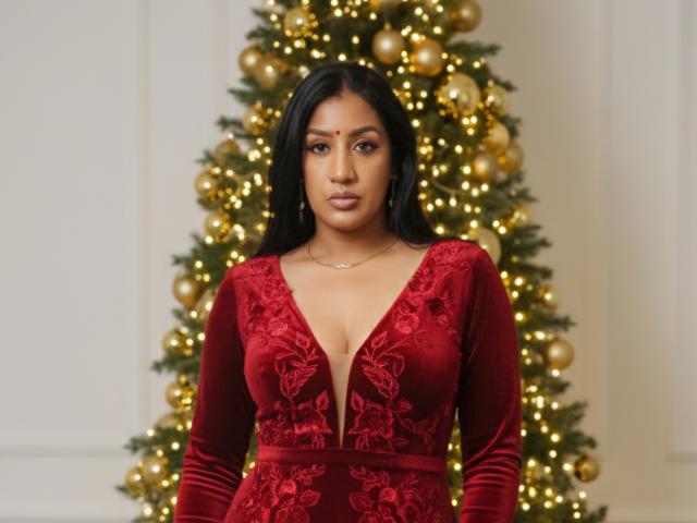 Nahirr with dark hair, red velvet dress with deep cleavage, floral embroidery, standing in front of a decorated Christmas tree, looking serious, medium skin, small nose, subtle makeup.