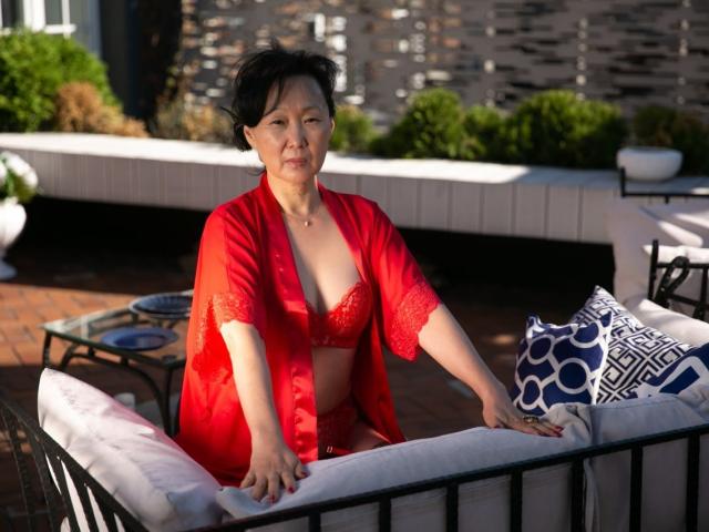 SandraMiy with short black hair, wearing red lace bra and sheer robe, sitting on a black metal patio chair with white cushions, looking at camera. Boobs visible.