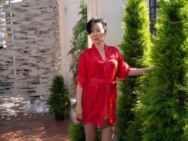 SandraMiy stands wearing a red sheer robe, exposing her small tits, against a green pine tree, with short black hair and a neutral expression.