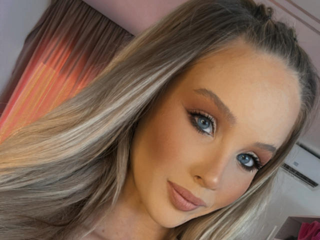 HeySiry has blue eyes, long blonde hair, and a cute face with subtle makeup, including pink lips and winged eyeliner. Her tits are not visible in this shot.