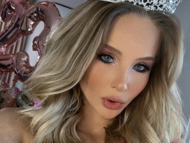 HeySiry has blonde wavy hair, blue eyes, and is wearing a silver crown, with pinkish lipstick and a slightly open mouth, looking directly at the camera.