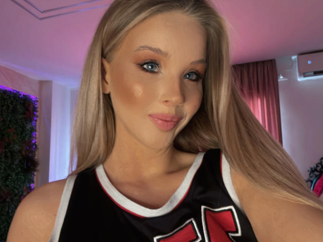 HeySiry with blonde hair, blue eyes, and pink lips, wearing a black and red basketball jersey, showing off her tits in a selfie.