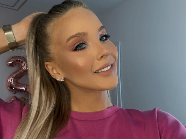 HeySiry has blonde ponytail, pink eyeshadow, shiny face, smiling, pink shirt, diamond earrings, wrist bracelet, looking at camera.