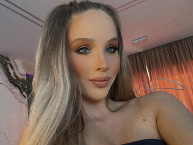 HeySiry has blue eyes, long blonde hair, and a cute smile, with subtle makeup and nude lipstick, showing her bare shoulders and a small mole on her chest.