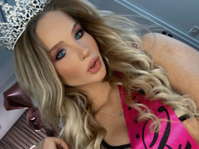 HeySiry has blonde curls, blue eyes, and a silver crown. She's wearing a pink sash and a black top, with her tits slightly visible. She's looking at the camera.