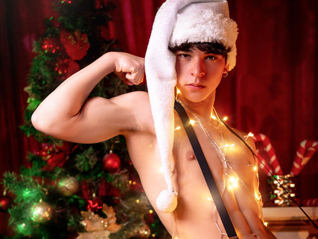 LukeHowlandy, zonder shirt, met gespierde arm, draagt een kerstmuts, kerstverlichting om de nek, bruin golvend haar, lichte huid, rode gordijn, kerstboom met versieringen, lolly, serieuze uitdrukking..