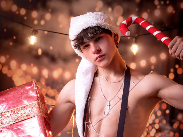 LukeHowlandy with a fluffy white Santa hat, holding a red candy cane and a shiny red gift, shirtless with a cross necklace, and string lights in the background.